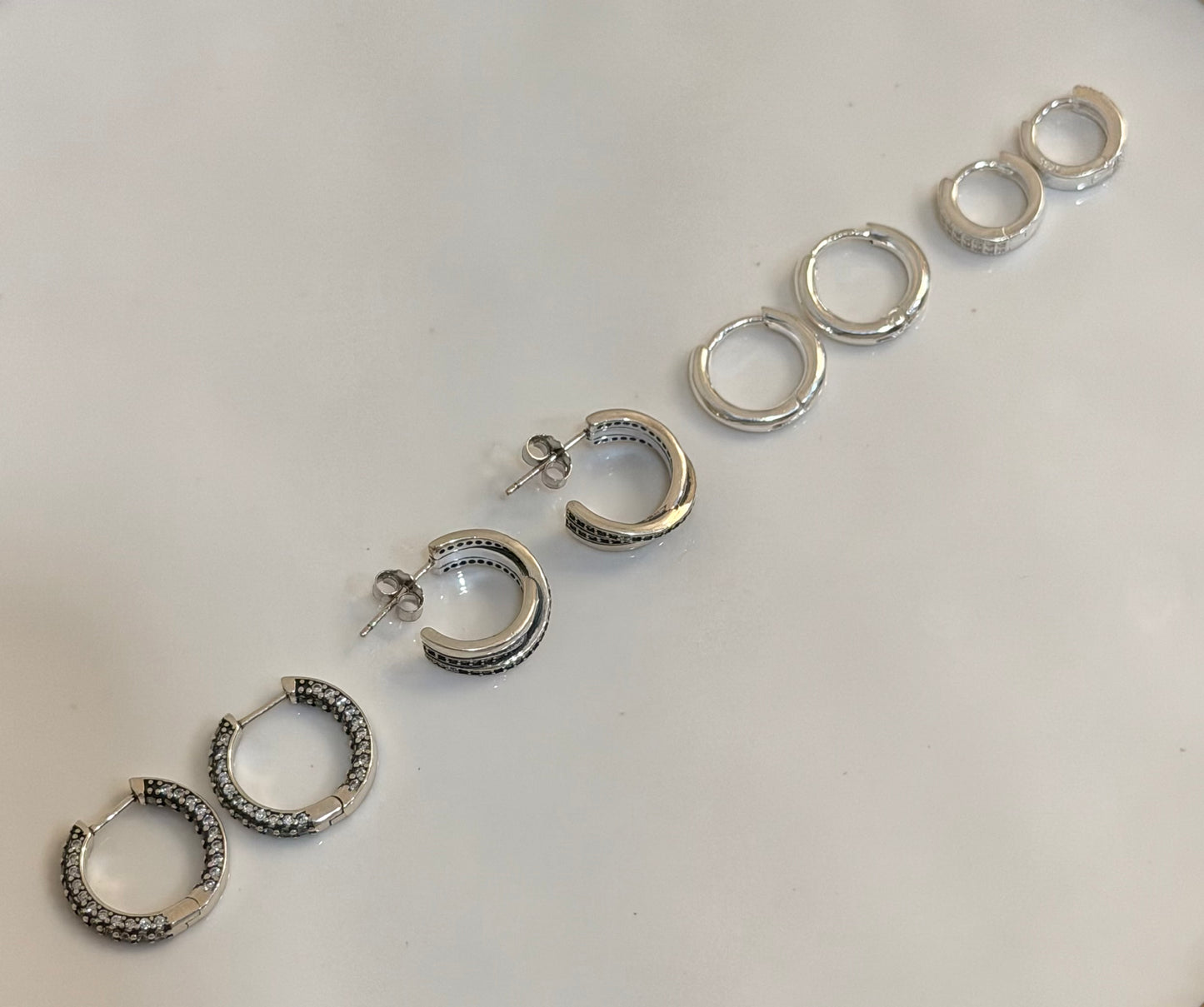 Six pairs of silver hoop earrings in various sizes and textures, arranged diagonally on a plain white surface.