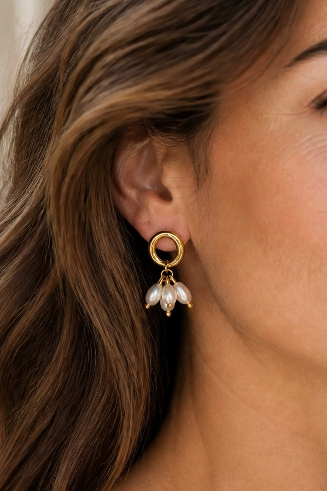 A close-up of a model's ear wearing a gold hoop earring with a dangling cluster of white pearl-like beads.