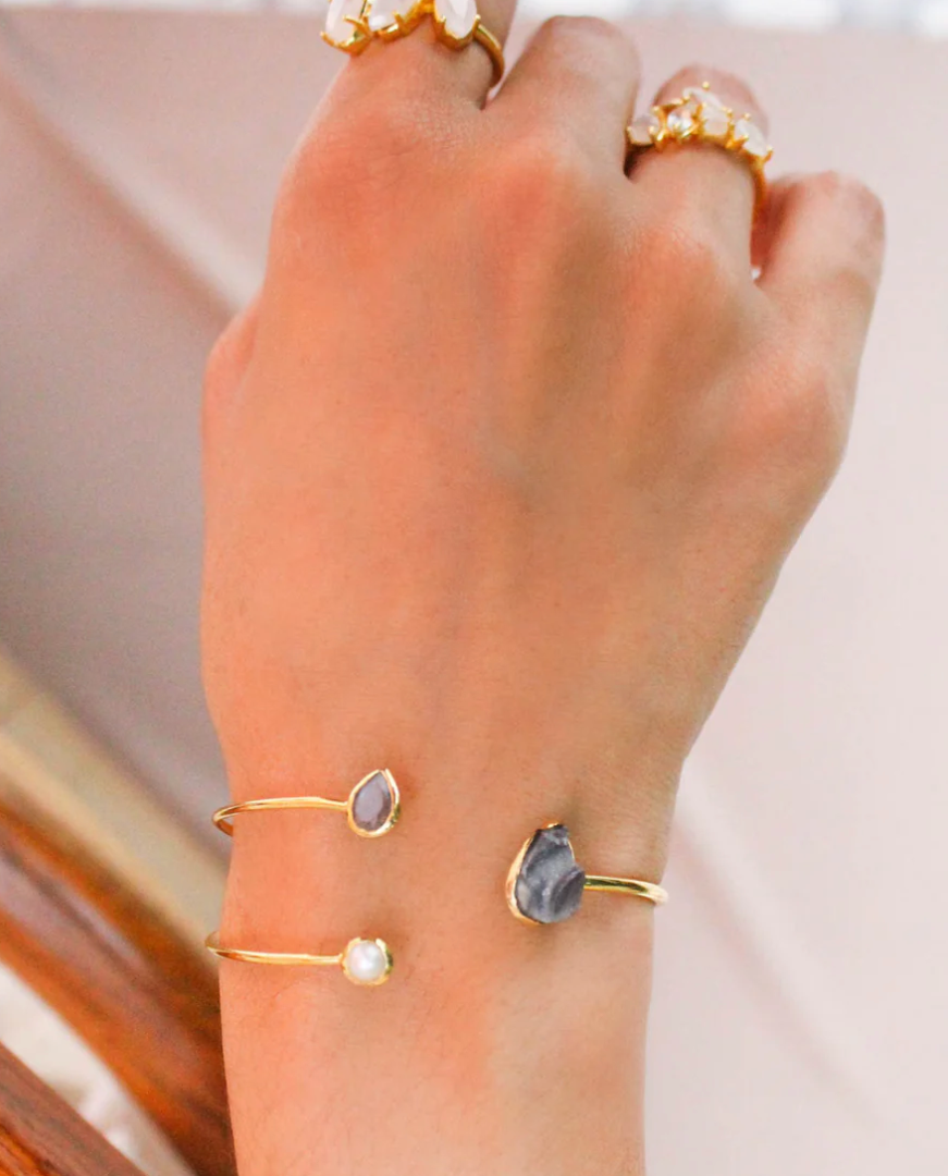 Stacked 18k gold-plated open cuffs on wrist with pearl, small grey stone, and teardrop blue stone ends; paired with gold floral rings.