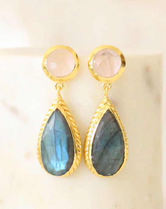 Pair of earrings with gold rims, pink gemstones, and blue teardrop stones on a beige background.