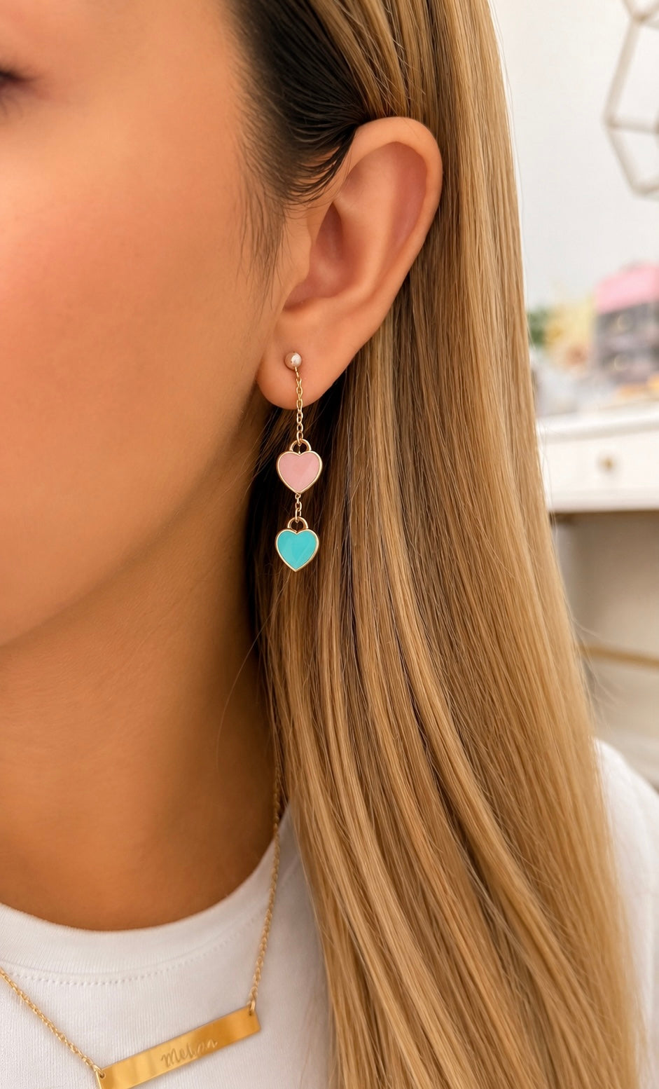 A close-up of a woman's ear wearing a gold drop earring with two dangling heart charms, one light pink and one light blue, paired with a white t-shirt.