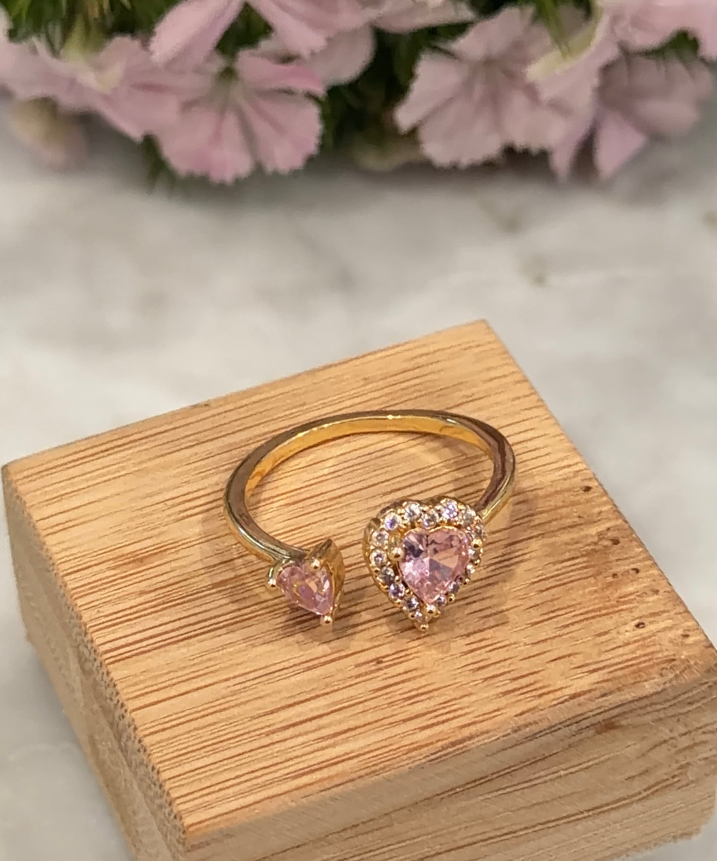 Gold ring with pink heart-shaped stones on a wooden box with flowers in the background