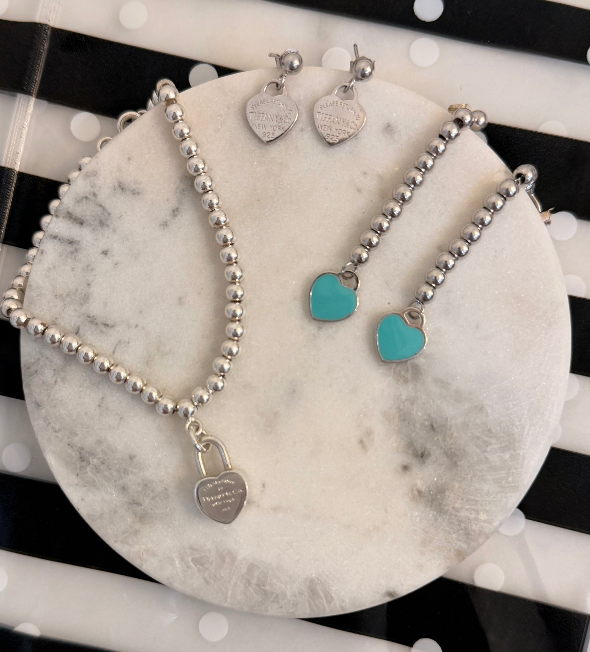 Silver beaded jewelry set with a heart-shaped padlock necklace, matching heart stud earrings, and heart-shaped drop earrings with turquoise heart charms, displayed on a round white marble tray.