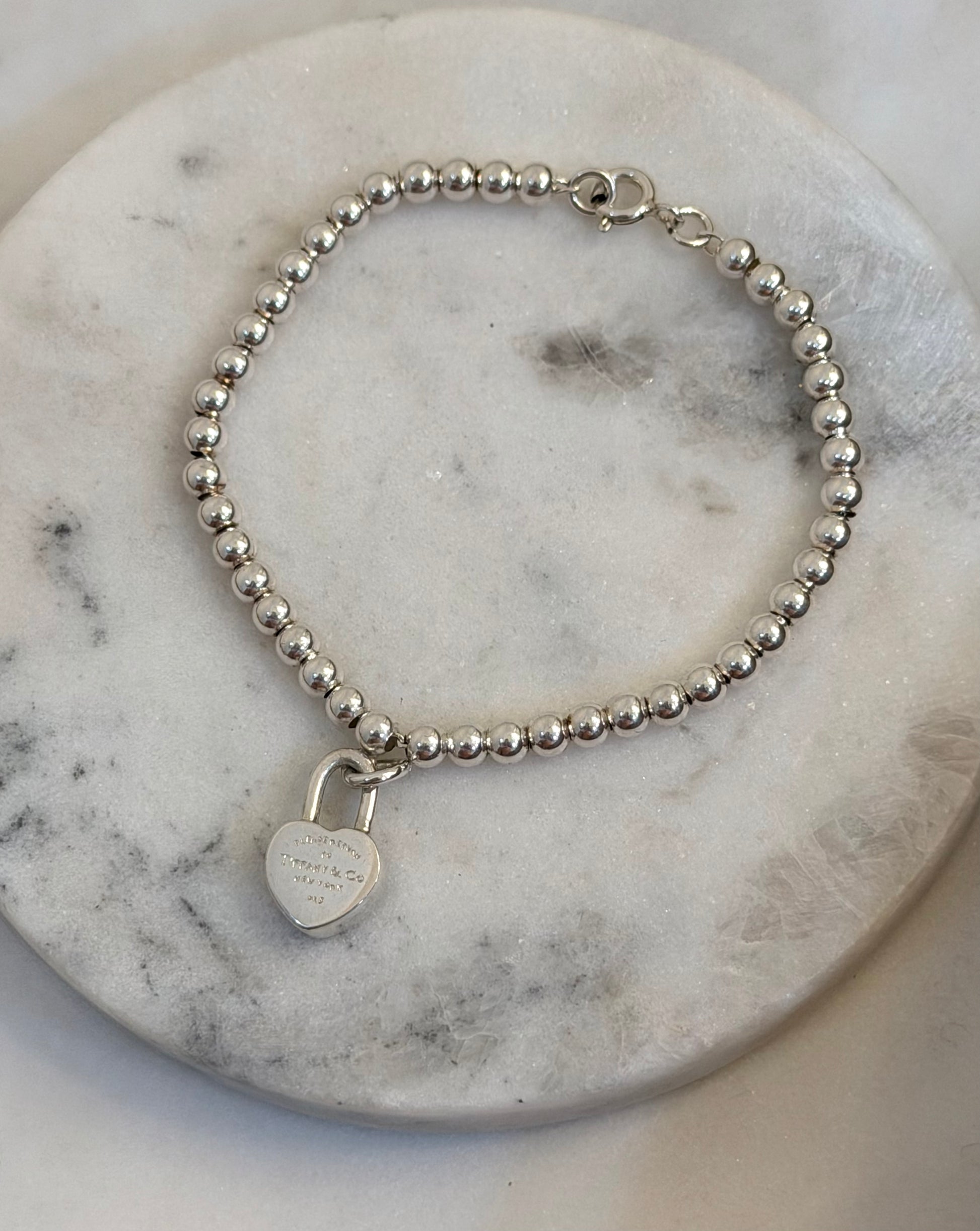 A silver beaded bracelet with a small heart-shaped padlock charm, displayed on a round white marble coaster.