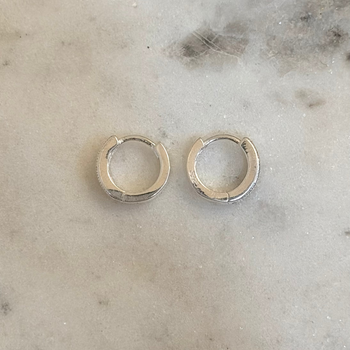 A pair of small silver huggie hoop earrings resting on a light gray marble surface.