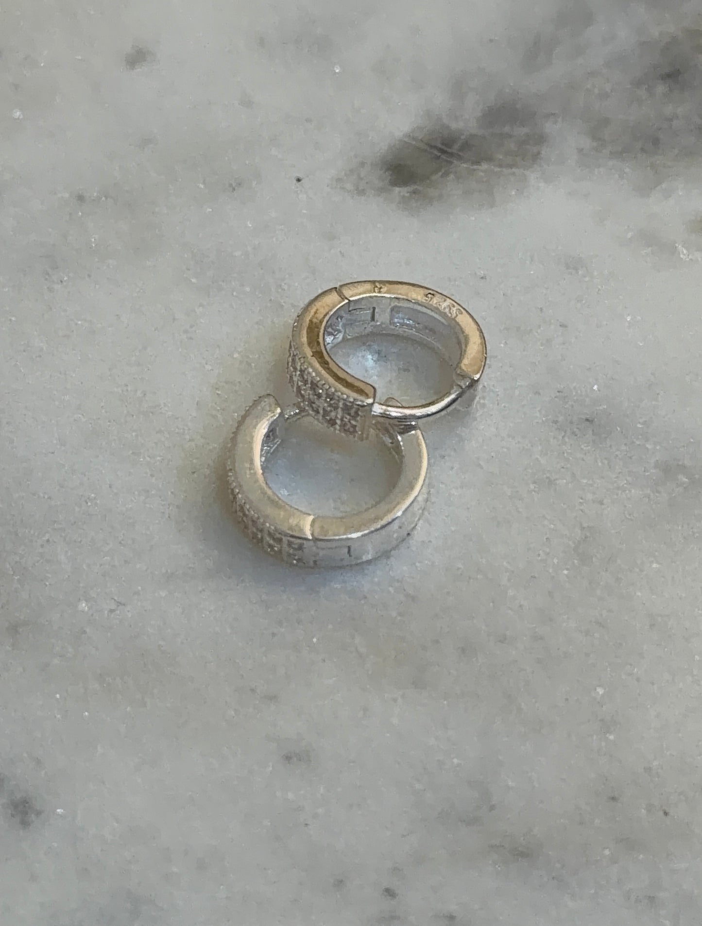 Two small silver huggie hoop earrings with textured, pavé-set stones, resting on a light gray marble surface.