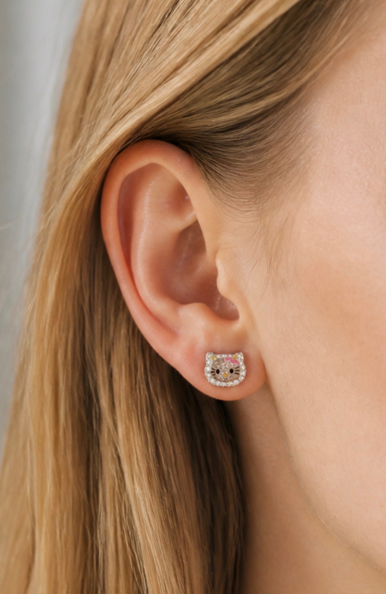A close-up of a model's ear wearing a small Hello Kitty stud earring with a pink and white face design.