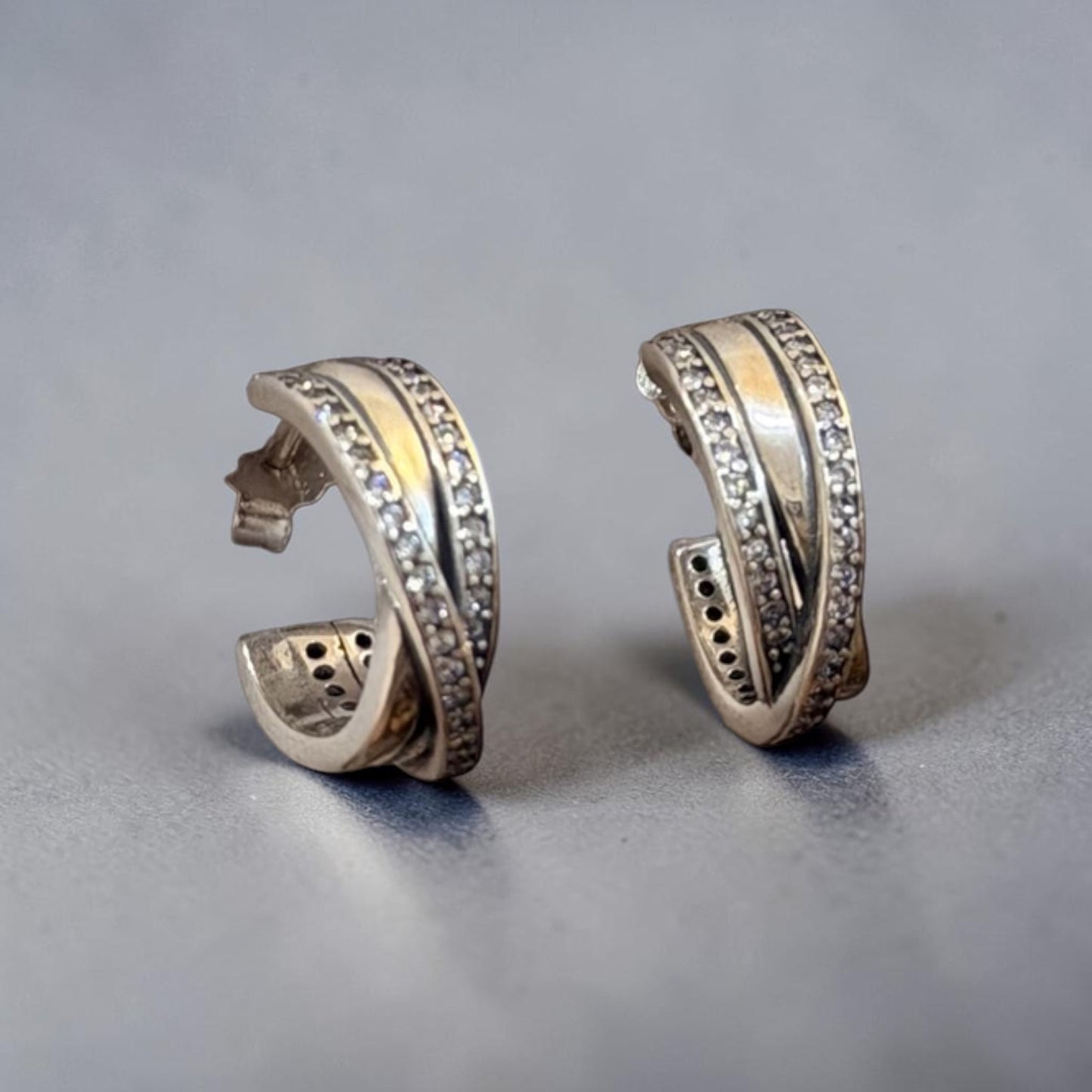 A pair of silver huggie hoop earrings with a twisted design, featuring one band set with small clear crystals and a polished yellow gold center stripe, displayed on a neutral gray surface.