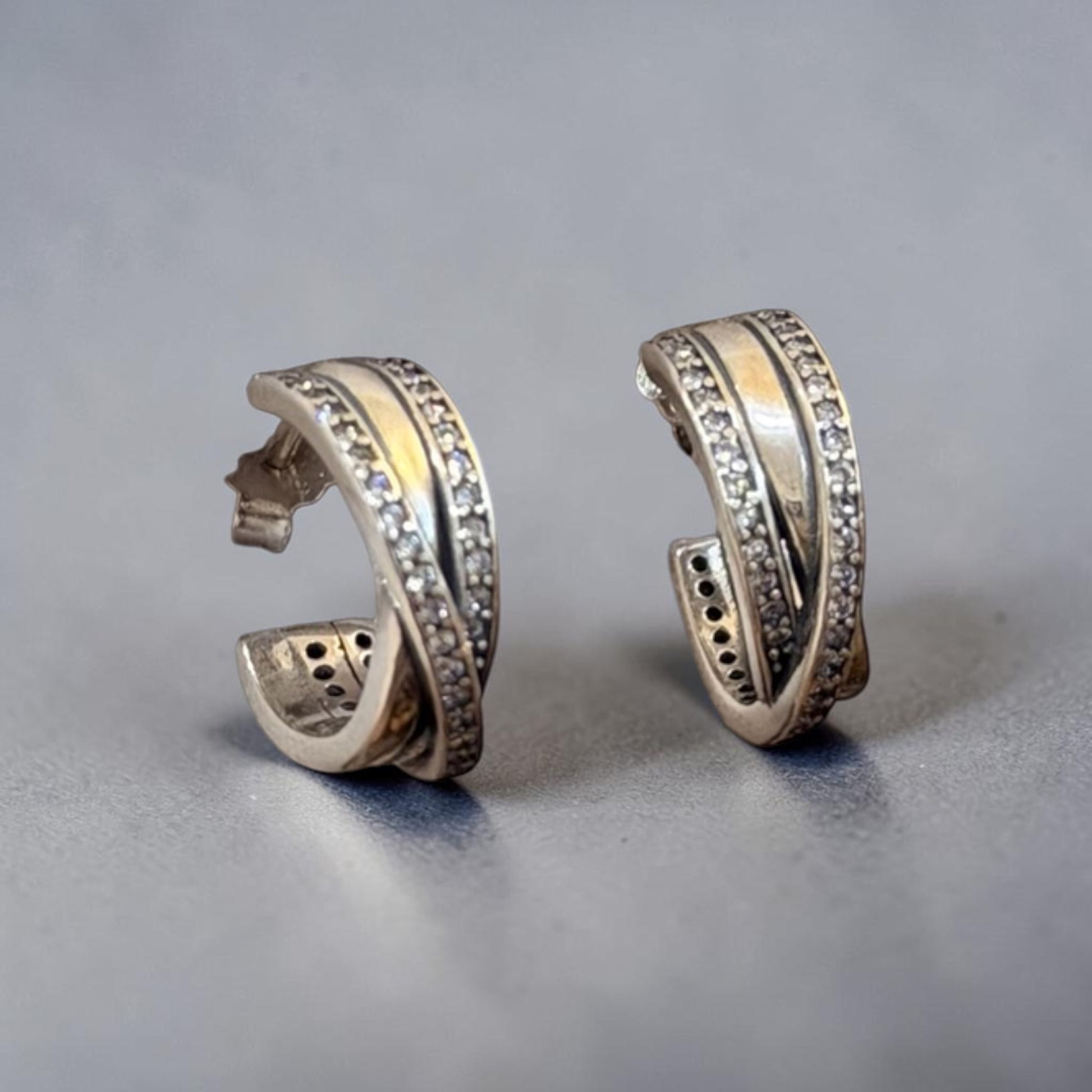 A pair of silver huggie hoop earrings with a twisted design, featuring one band set with small clear crystals and a polished yellow gold center stripe, displayed on a neutral gray surface.