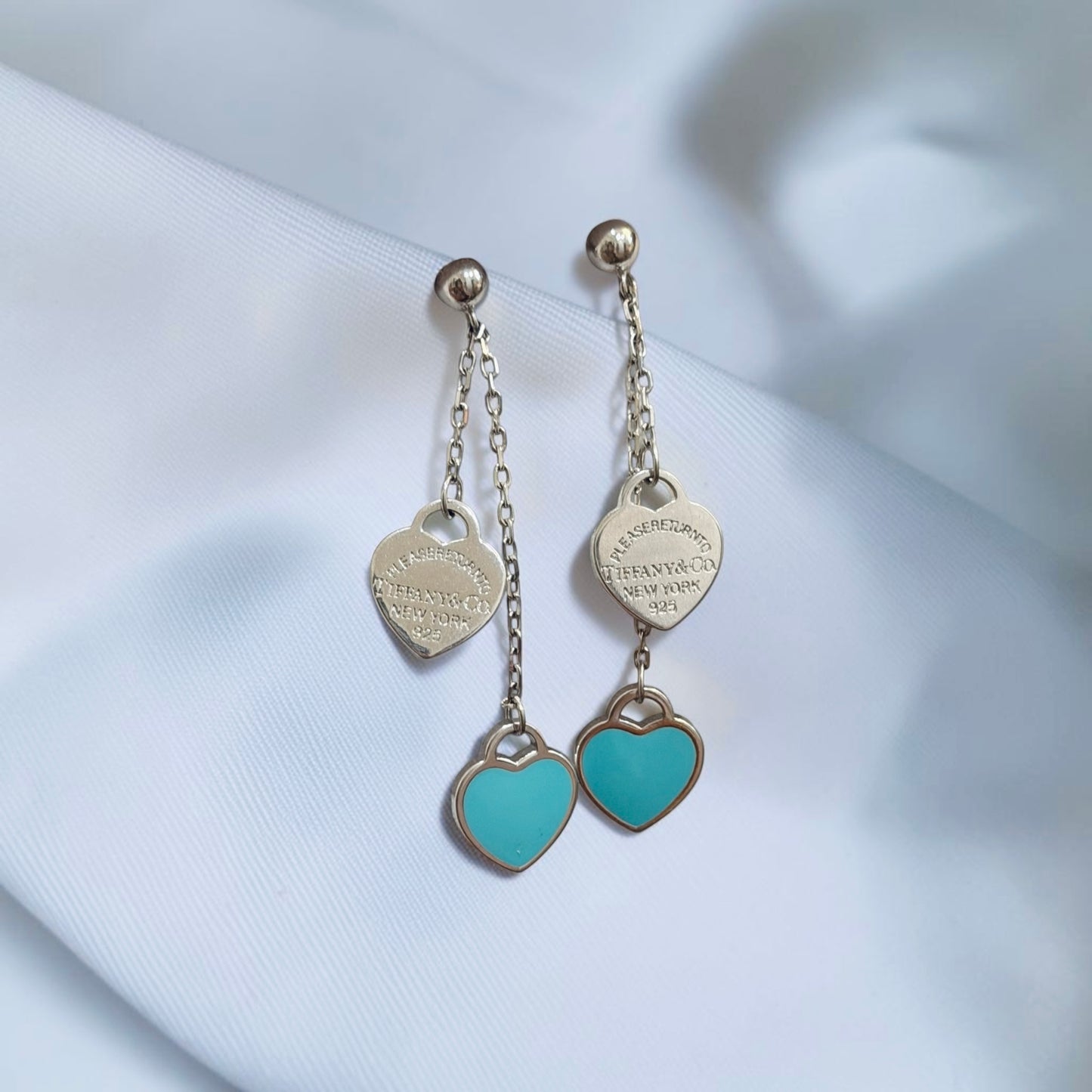 A pair of silver-toned dangle earrings with heart-shaped charms, displayed on a soft white fabric background. Each earring features a small silver ball stud, a chain, a heart-shaped engraved charm, and a larger dangling turquoise heart charm.