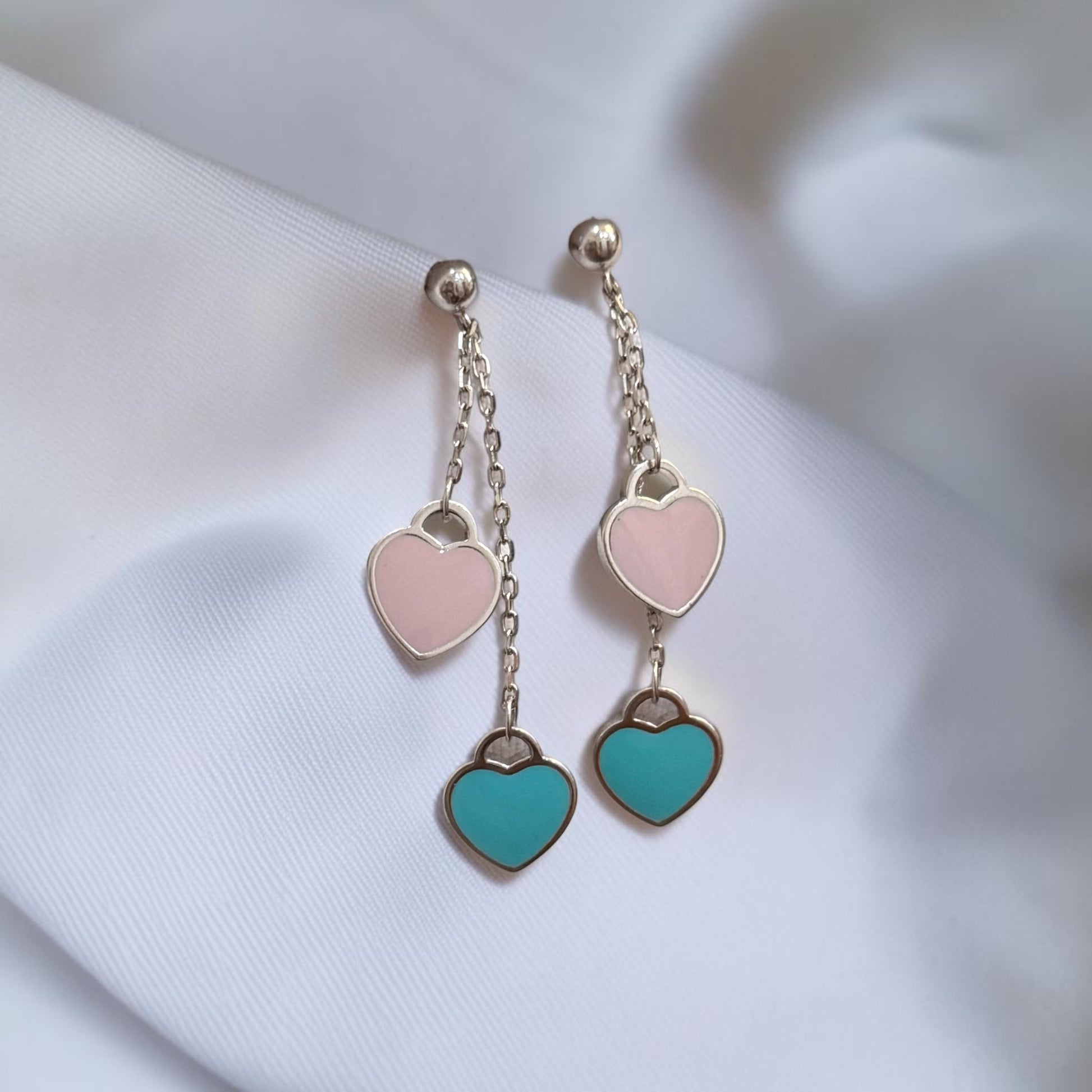 A pair of dangling heart earrings with silver-tone chains, featuring a light pink heart and a turquoise heart, displayed on a soft white fabric background.
