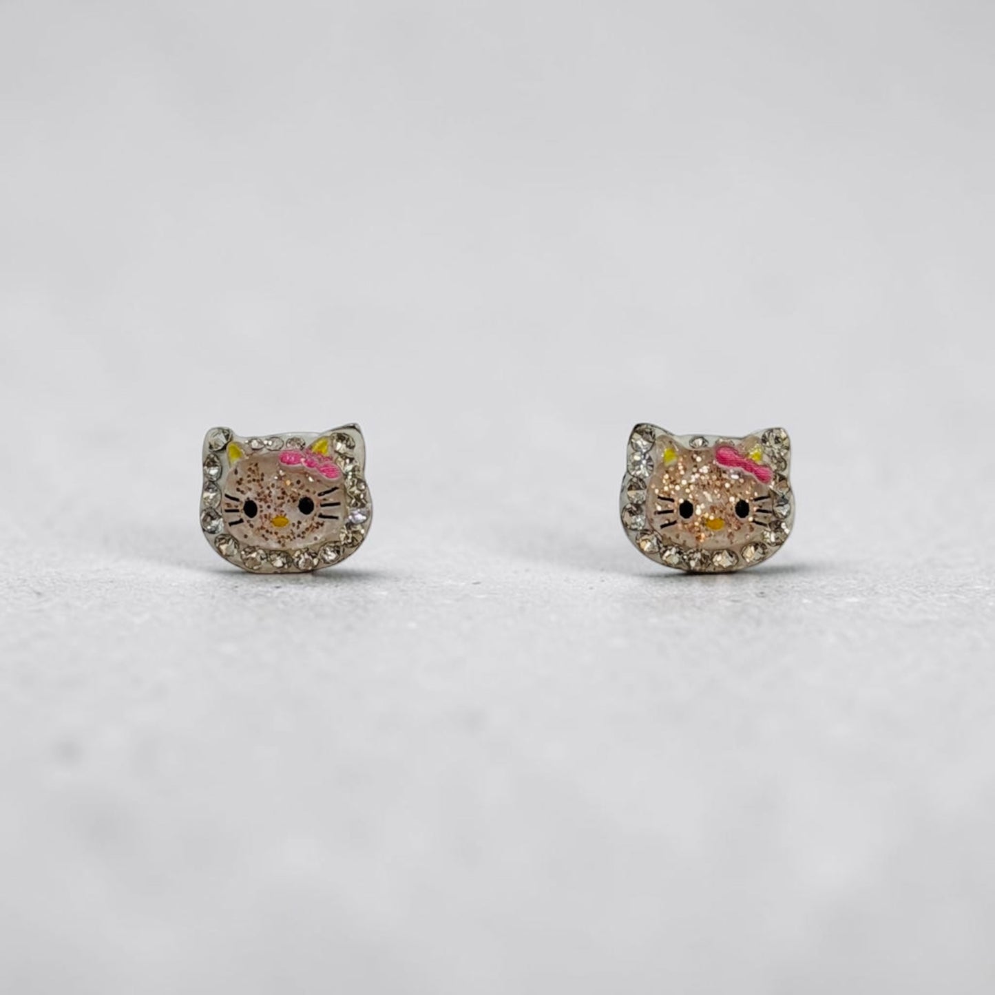 A pair of silver stud earrings shaped like Hello Kitty's face, embellished with clear rhinestones and featuring pink and yellow enamel details, set against a light gray background.