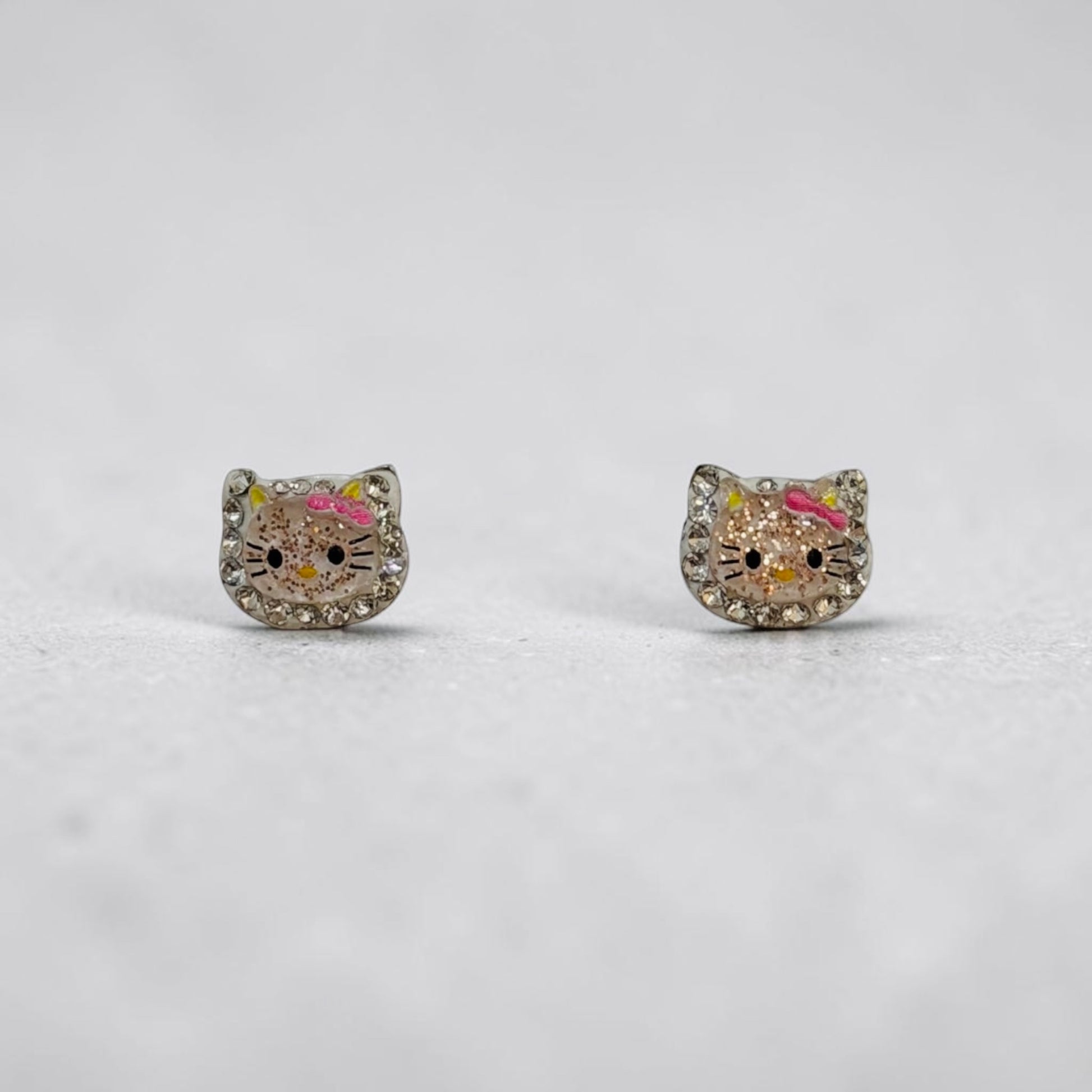 A pair of silver stud earrings shaped like Hello Kitty's face, embellished with clear rhinestones and featuring pink and yellow enamel details, set against a light gray background.