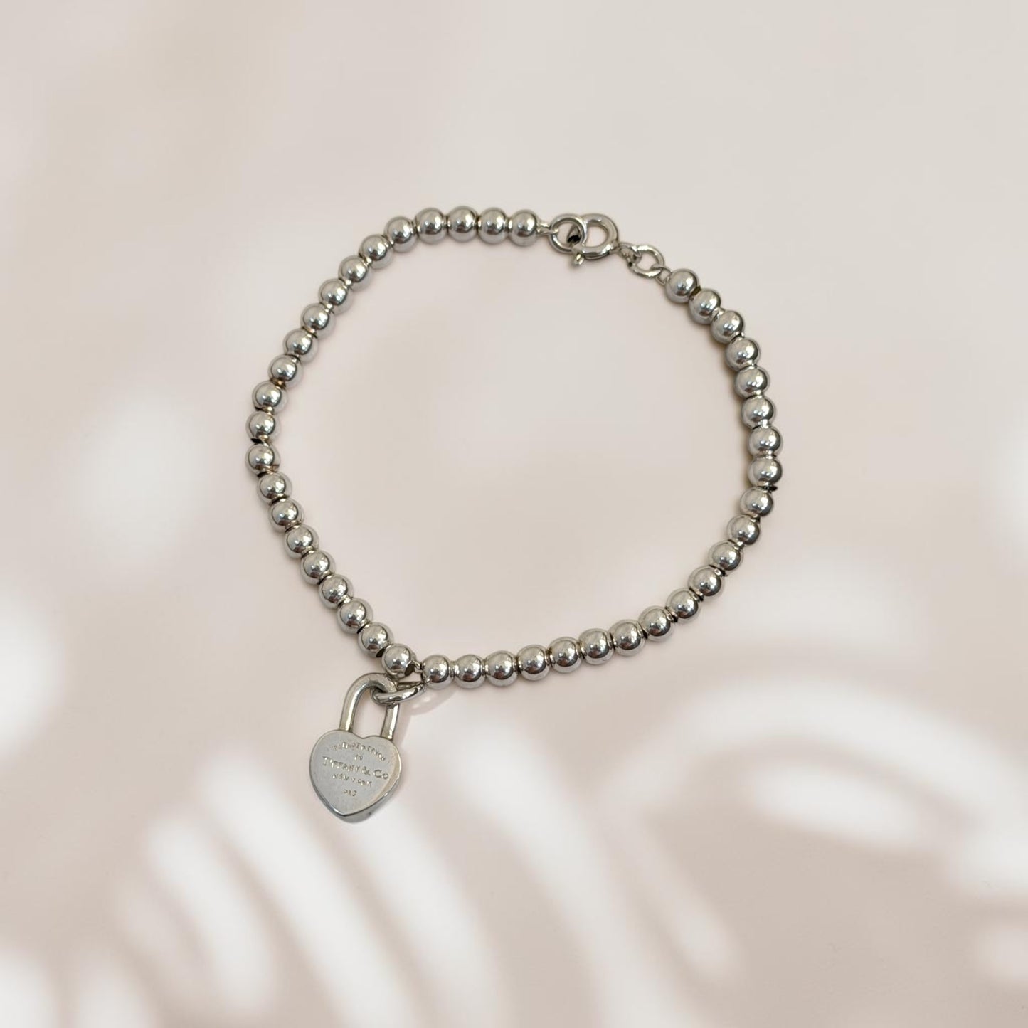 A silver beaded bracelet with a small heart-shaped padlock charm, displayed on a soft beige background with subtle leaf shadows.