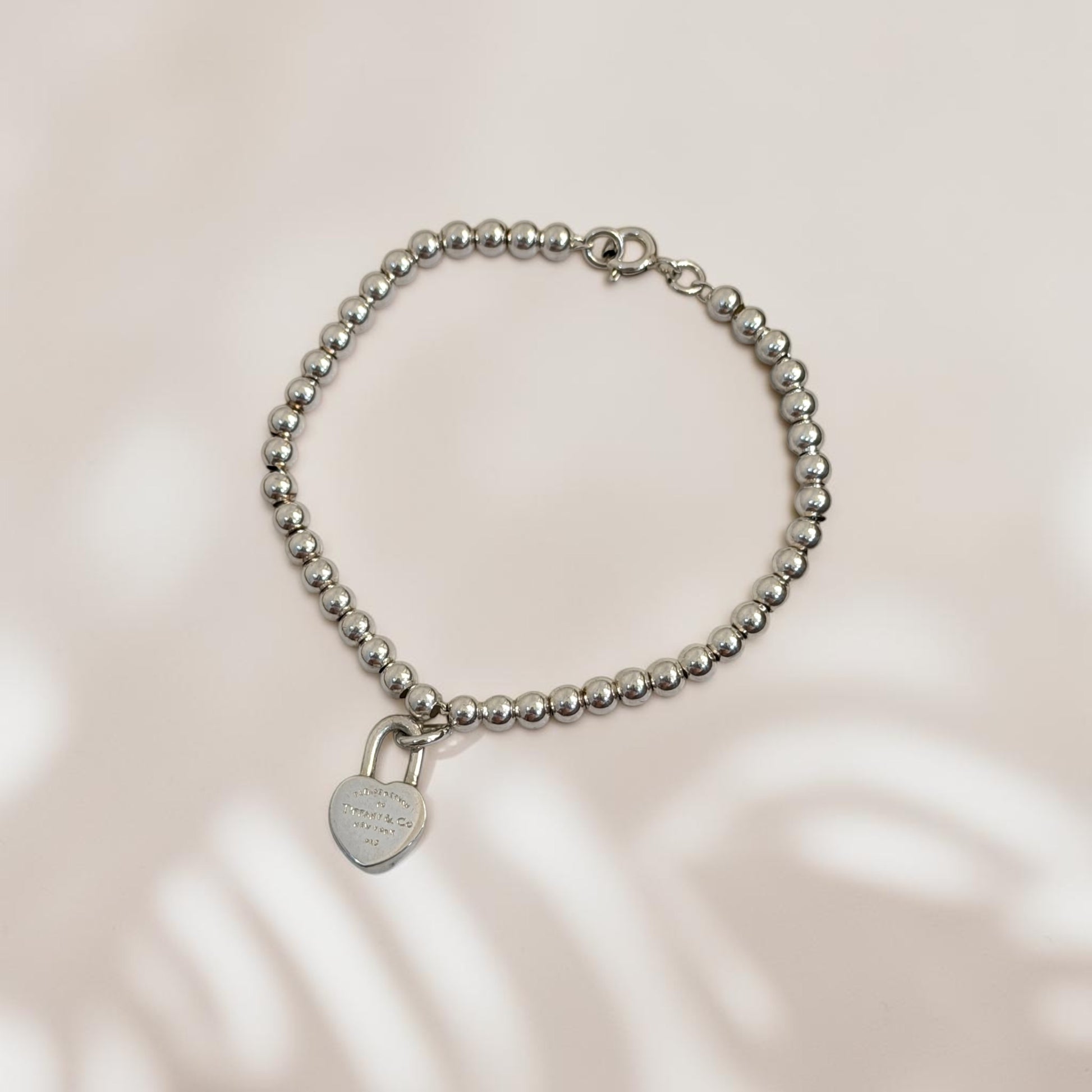 A silver beaded bracelet with a small heart-shaped padlock charm, displayed on a soft beige background with subtle leaf shadows.