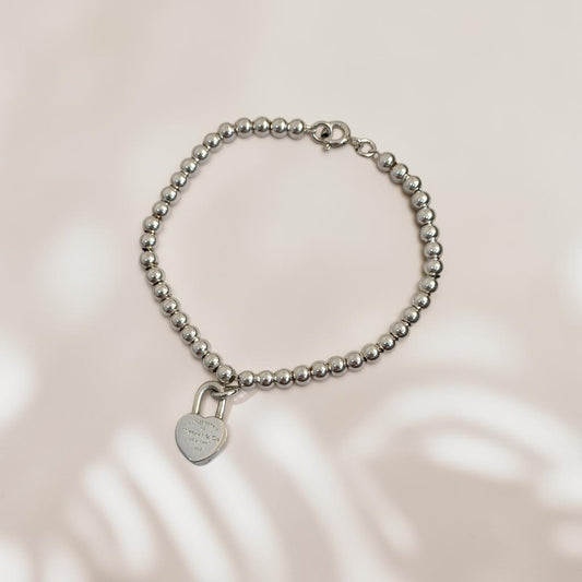 A silver beaded bracelet with a small heart-shaped padlock charm, displayed on a soft beige background with subtle leaf shadows.